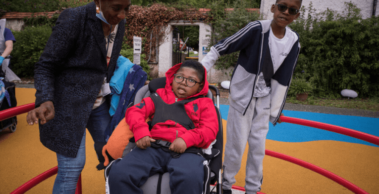 A young boy is on an accessible roundabout in his wheelchair, with his brother and carer